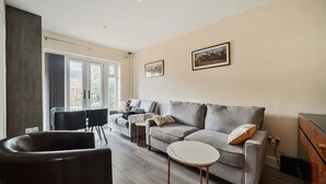 A modern living room with a grey sofa, a coffee table, and a black chair. There is a glass table with chairs, a window with blinds, and a painting on the wall. - Close to Windsor, Heathrow and central London — sleeps nine (Berkshire)
