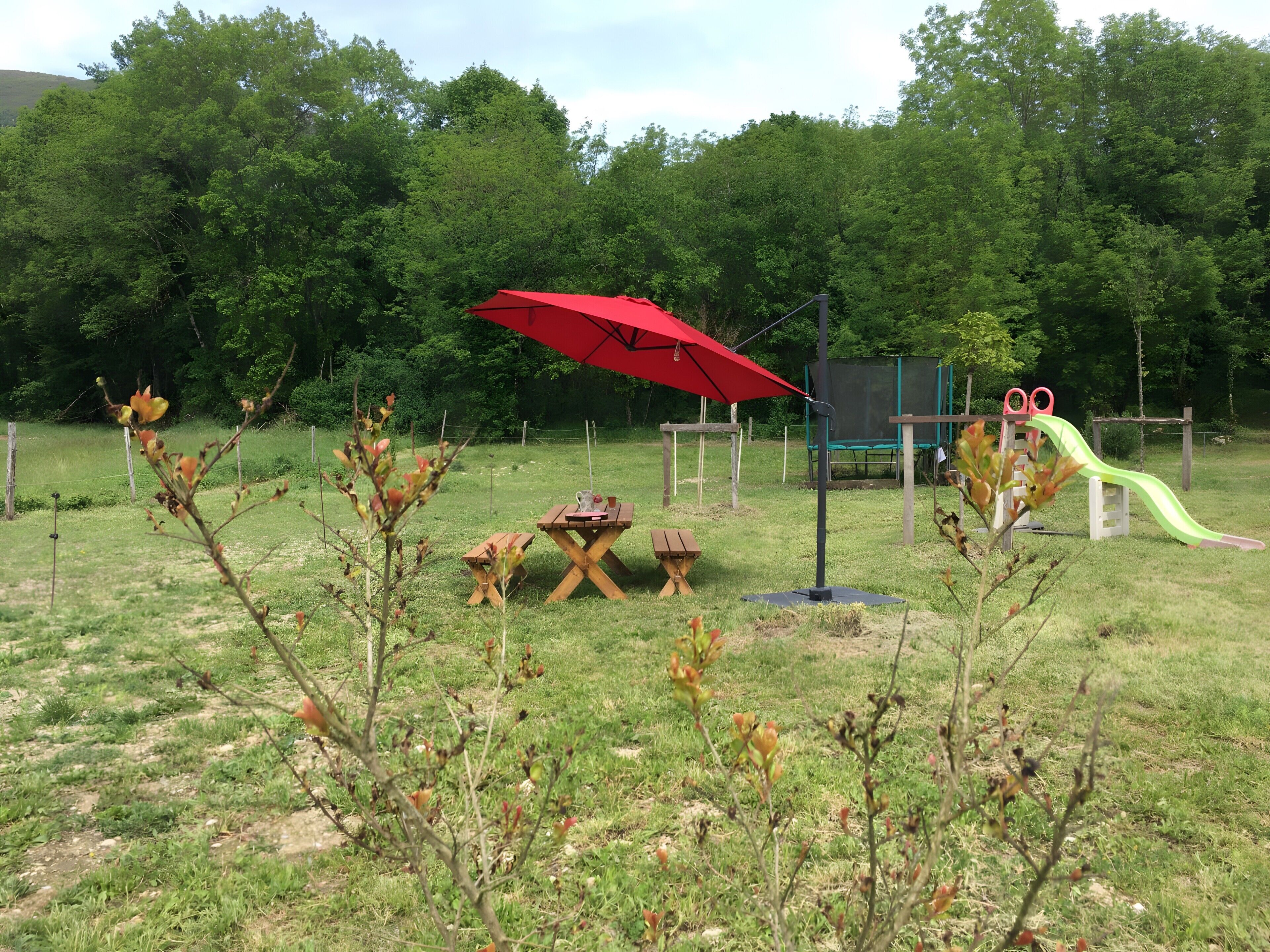 Un espace extérieur verdoyant avec des tables de pique-nique, un parasol rouge et une aire de jeux dotée d’un toboggan.