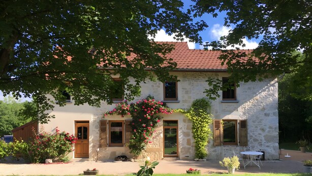Ein zweigeschossiges Haus aus Stein mit rotem Ziegeldach, einer Haustür und einer kleinen Terrasse mit Tisch und Stühlen.