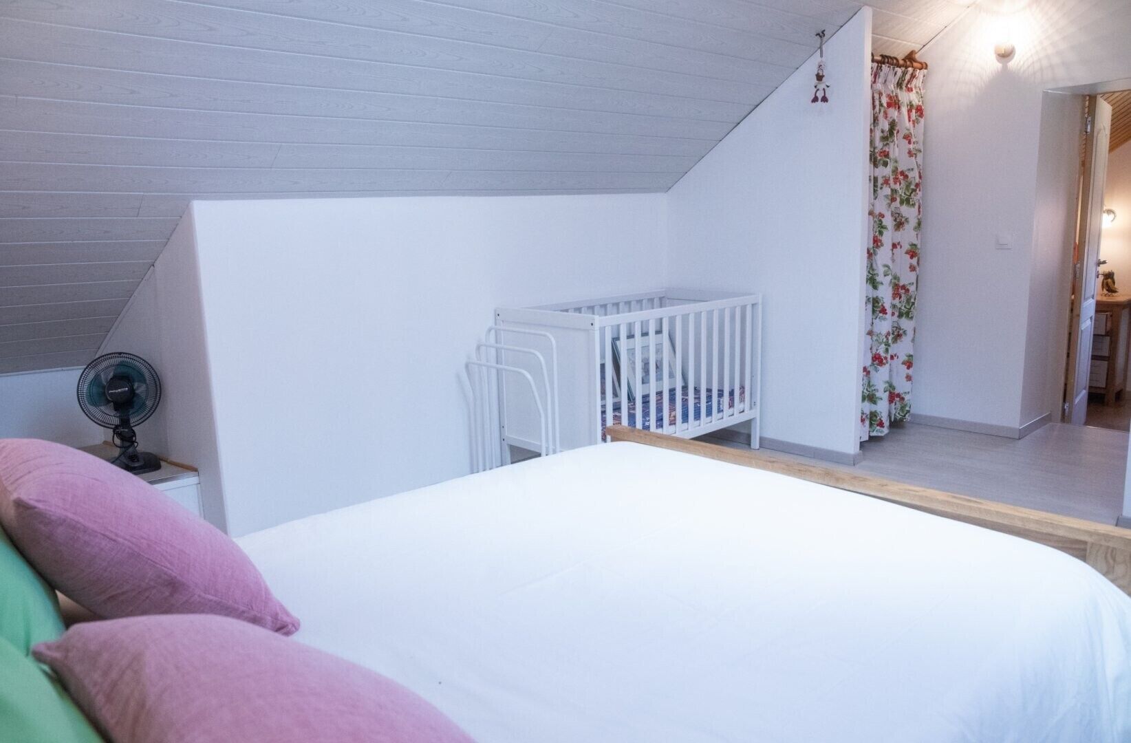 A bedroom with a white bed, a crib, a fan, and a staircase leading to an upper level.