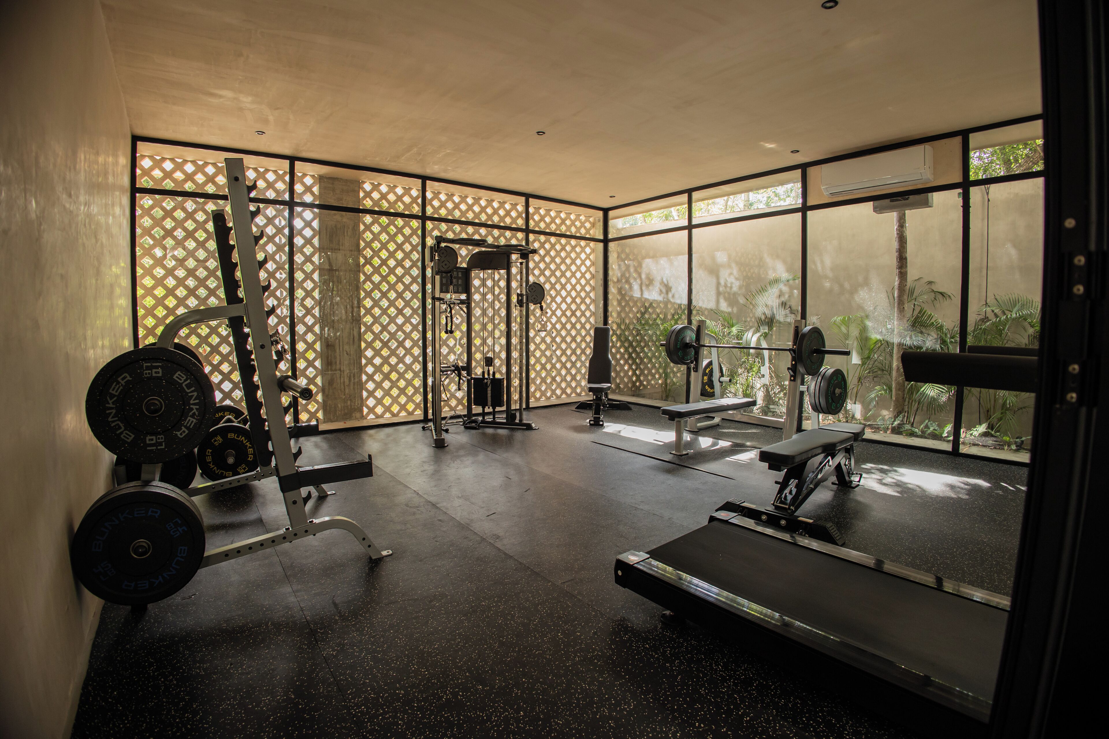 Un gimnasio con equipo de levantamiento de pesas, cinta de correr y amplias ventanas con vistas a zonas verdes.