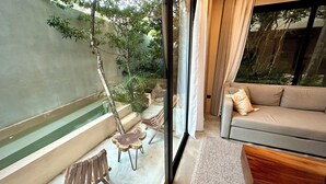 A modern living room with a sofa, a small wooden table, and chairs. There is a glass wall with a view of greenery outside. - Modern duplex with private pool + 2 bicycles / 7 minutes from Tulum Beach (Tulum)