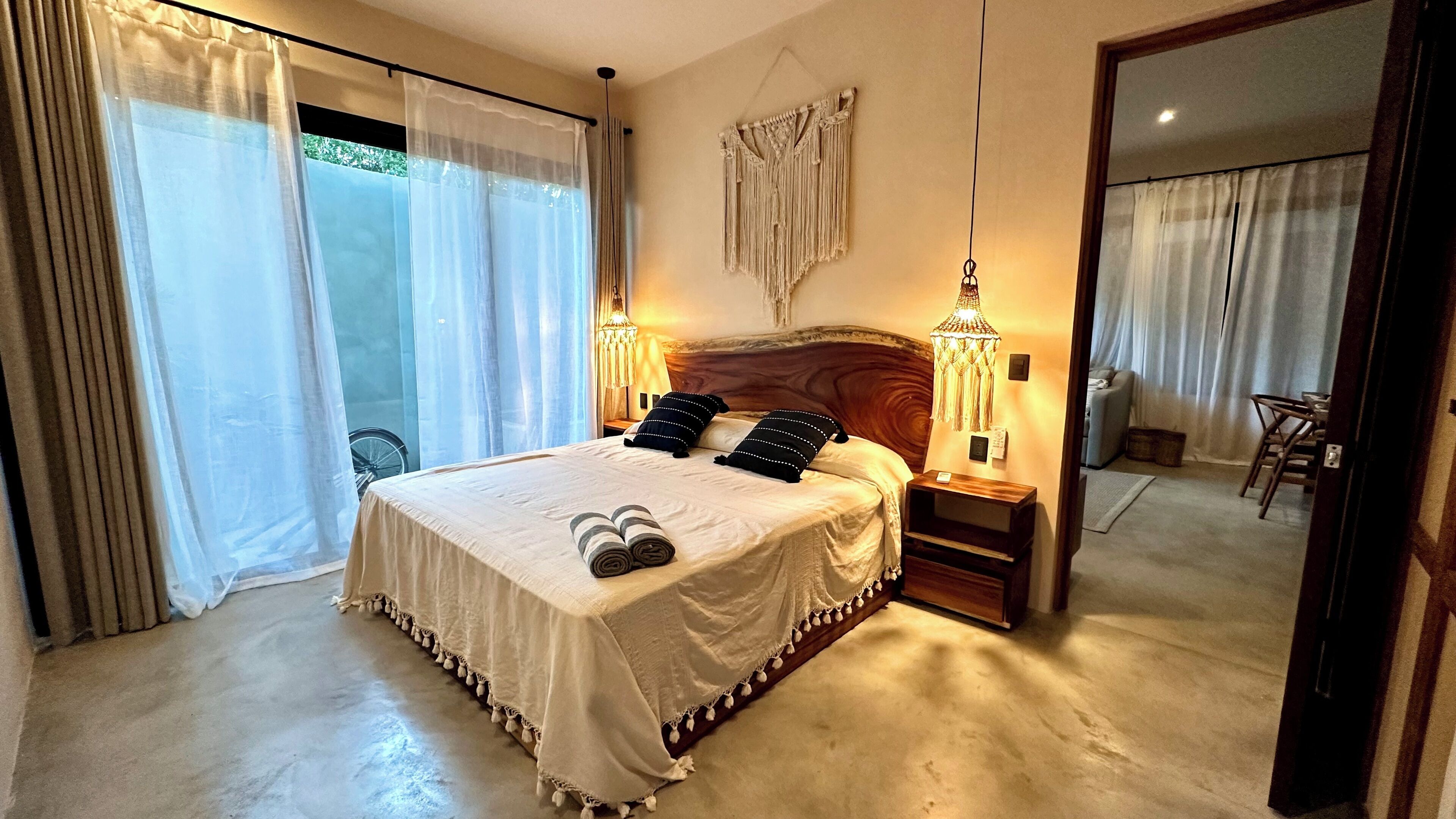 Habitación de hotel con una cama grande, dos lámparas de noche, un espejo y vistas al exterior a través de cortinas translúcidas.