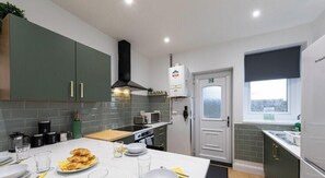 A modern kitchen with grey cabinets, a microwave, a stove, and a dining area set with plates and glasses. - Luke Stays - Grace Street (Tyne and Wear)
