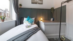 A neatly arranged hotel room with a bed, a bedside table with a lamp, a window with curtains, and a view of the outside. - Luke Stays - Grace Street (Tyne and Wear)