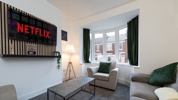 A living room with a Netflix sign, a lamp, a sofa, a chair, and a window with curtains. - Luke Stays - Grace Street (Tyne and Wear)