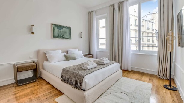 Ein modernes Hotelzimmer mit einem großen Bett, einem Nachttisch, einer Stehlampe und Blick auf Gebäude durch das Fenster.