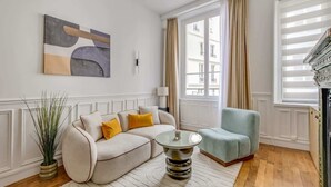 Ein modernes Wohnzimmer mit einer Couch, einem runden Couchtisch und einem Sessel. An der Wand hängt ein Gemälde, eine Pflanze steht dort und ein Fenster ist mit Vorhängen drapiert.