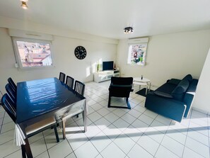 A living room with a dining table, chairs, a sofa, a TV, and a clock on the wall. - Comfortable stay in Gérardmer, near the lake (Gérardmer)