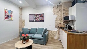 Una sala de estar moderna con un sofá color turquesa, una pequeña mesa redonda con una planta, y una zona de cocina con una pared de piedra y armarios blancos.