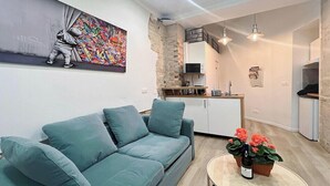 Una sala de estar moderna con un sofá azul aguamarina, una pequeña mesa de centro con una planta, y una zona de cocina con una pared de piedra y armarios blancos.