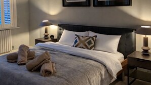 Un dormitorio con una cama grande, dos mesitas de noche con lámparas, una ventana con persianas y dos cuadros en la pared.