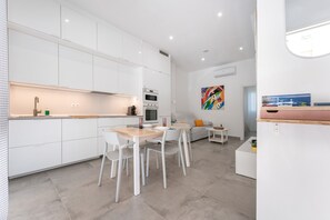 Una cocina moderna con armarios blancos, un comedor con mesa de madera y sillas blancas, y una sala de estar con un sofá y un cuadro de colores.