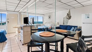 Quarto moderno com área de jantar, sofá, televisão e vista para o mar.