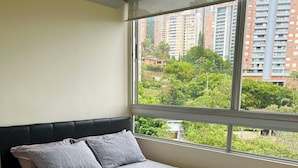 Une chambre avec un grand lit, une fenêtre donnant sur des immeubles de grande hauteur et sur un espace verdoyant.