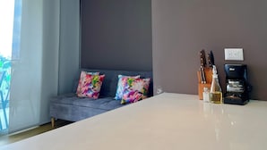 Une pièce moderne avec une grande table, deux lampes suspendues, un canapé agrémenté de coussins colorés, une machine à café et un bloc à couteaux.