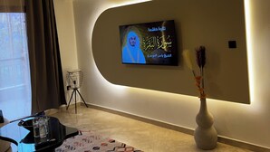 Ein modernes Wohnzimmer mit einem flachbildfernseher an einer gewölbten Wand, einer Stehlampe, einem gemusterten Teppich und einer Vase mit getrockneten Blumen.