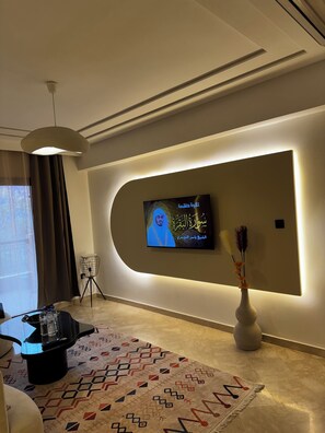 Ein modernes Wohnzimmer mit einem flachbildfernseher an einer gewölbten Wand, einer Stehlampe, einem gemusterten Teppich und einer Vase mit getrockneten Blumen.