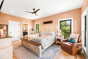 Ein Schlafzimmer mit Bett, Sitzbank, Kommode, Stuhl, Nachttisch, Lampe, Deckenventilator, Fenster und Tür.