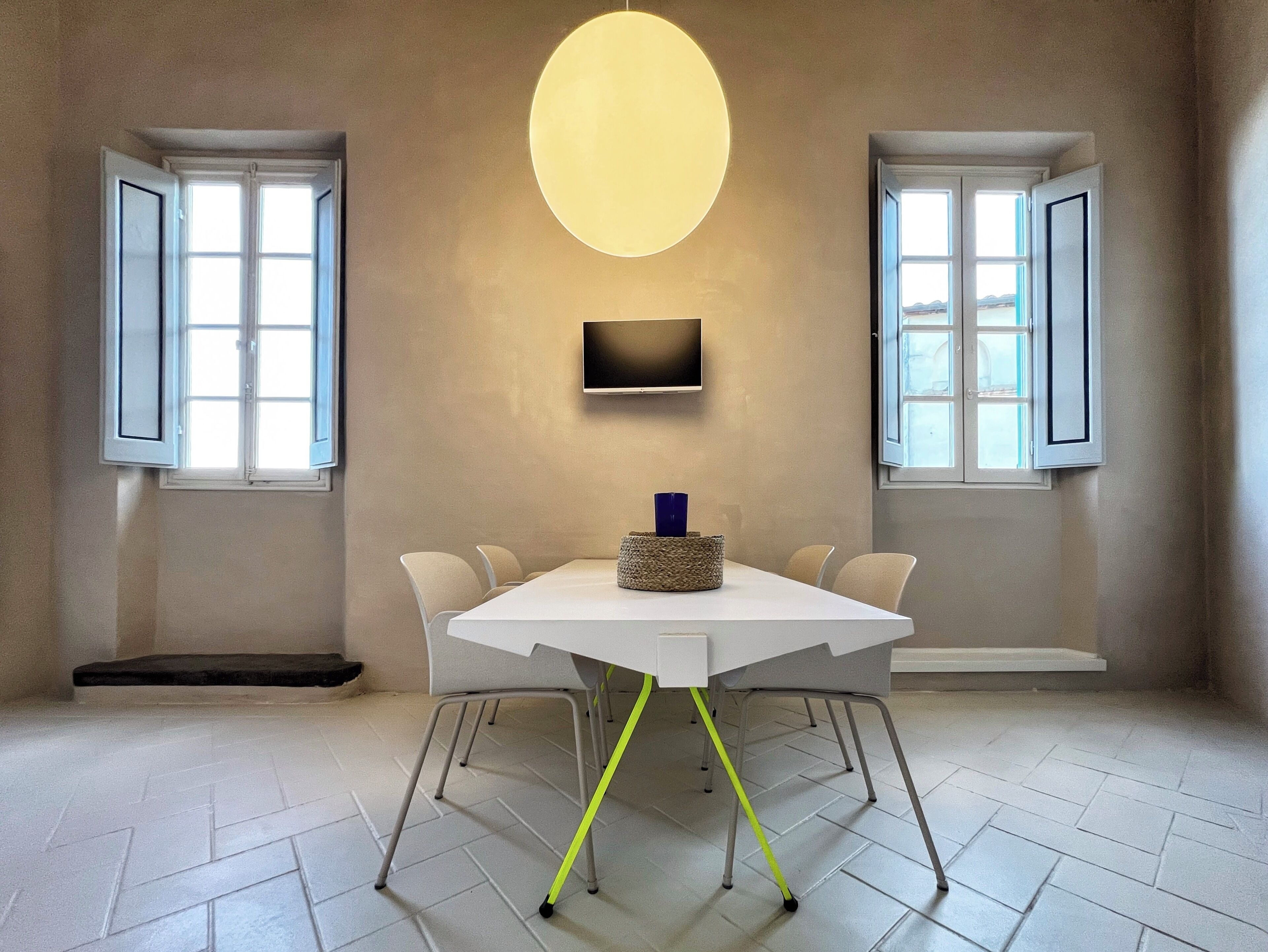 Una habitación moderna con una mesa blanca, cuatro sillas, un televisor de pantalla plana y dos ventanas con contraventanas abiertas.