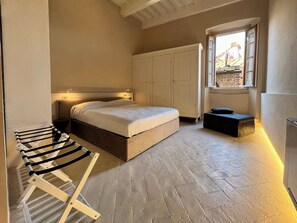Un dormitorio con cama, armario, banca, tendedero y ventana con vista a un edificio.
