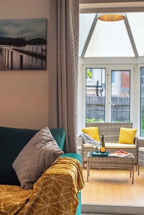 Un salon chaleureux avec un canapé, un fauteuil en osier et une table sur laquelle se trouvent une bouteille et de la nourriture.