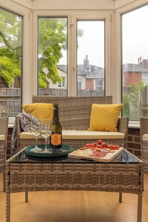Un canapé en osier avec des coussins jaunes, une table en verre avec une bouteille et des verres, et un plateau avec de la nourriture.
