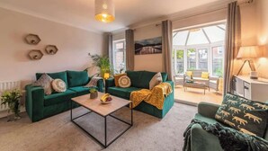 A living room with a green sofa, a coffee table, and a lamp. There is a window with curtains and a view of the outside. - 3BR Luxury Chic Spacious House w/Parking -Sleeps 7 (Southampton)