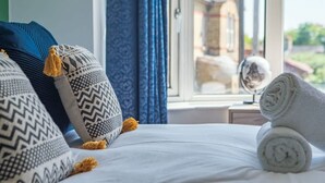 Cama com almofadas estampadas, toalha enrolada e vista de um edifício pela janela.