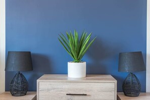 A room with a blue wall, a wooden nightstand with a drawer, a white pot with a green plant, and two black lamps with woven bases. - 3-Bed Family House with Garden, Garage & Free Parking, Southampton (Southampton)