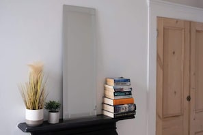 Eine Ecke mit weißer Wand, einem Spiegel, einem schwarzen Regal mit Pflanze und Büchern sowie einer Holztür.
