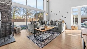 Ein geräumiges Wohnzimmer mit großem Fenster, einer Steinwand, einem grauen Sofa, einem Holztisch und einer schwarzen Metallstehlampe.