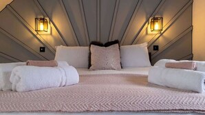 Une chambre d’hôtel moderne avec un plafond au design géométrique, deux lampes fixées au mur, un grand lit surmonté d’un tapis rose à motifs, et des serviettes soigneusement pliées.