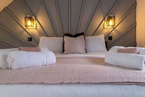 Ein modernes Hotelzimmer mit einem geometrischen Deckenmuster, zwei Wandleuchten, einem großen Bett und einem rosa Textilteppich. Ordentlich gefaltete Handtücher vervollständigen das Bild.