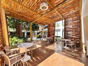 Un espace extérieur aménagé en terrasse, avec un sol en bois, des tables et des chaises, entouré de plantes et doté d’un plafond en bambou.