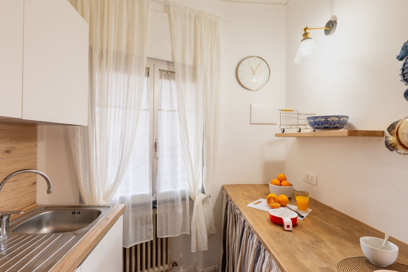 A kitchen with a sink, a clock, a fruit bowl, and a cutting board.