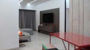 A modern hotel room with a grey sofa, a red table, a flat-screen TV, and a grey curtain.