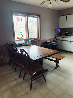 Un coin repas avec une table et des chaises en bois, une fenêtre avec des stores et un espace cuisine équipé de placards et d’appareils électroménagers.