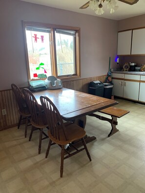 A dining area with a wooden table and chairs, a window with blinds, a ceiling fan, and a kitchen area with cabinets and appliances.