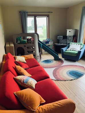 A room with a red sofa, a blue chair, a playpen, and a window with curtains.