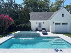 Una piscina rectangular con borde de piedra, una silla de descanso y una planta en maceta cerca de una casa blanca con garaje.