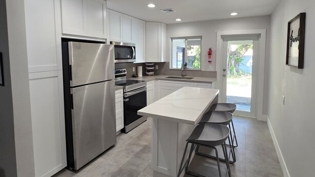 A modern kitchen with white cabinetry, stainless steel appliances, and a central island with bar stools. - Cozy 2BD/1BA Near Beach + Fast I-95 Access! (Hallandale Beach)