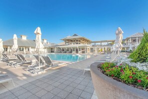 Un espace piscine avec des chaises longues et des parasols, un ciel dégagé et, au loin, un bâtiment à l’architecture moderne.