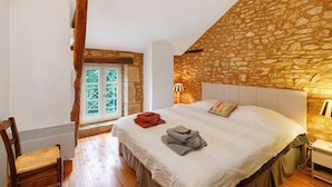 A bedroom with a stone wall, a bed with a white headboard, a wooden chair, a radiator, and a window with a view. - Gîte “La Gariotte” with Shared Pool and Jacuzzi (Montcabrier)