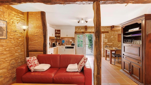 A cozy living area with a red leather sofa, wooden beams, stone walls, and a view of the kitchen and outdoor space. - Gîte “La Gariotte” with Shared Pool and Jacuzzi (Montcabrier)