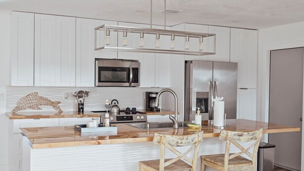 Een moderne keuken met witte kastjes, een roestvrijstalen koelkast, een magnetron en een ingebouwde oven. Er is een houten kookeiland met een spoelbak en twee houten stoelen.
