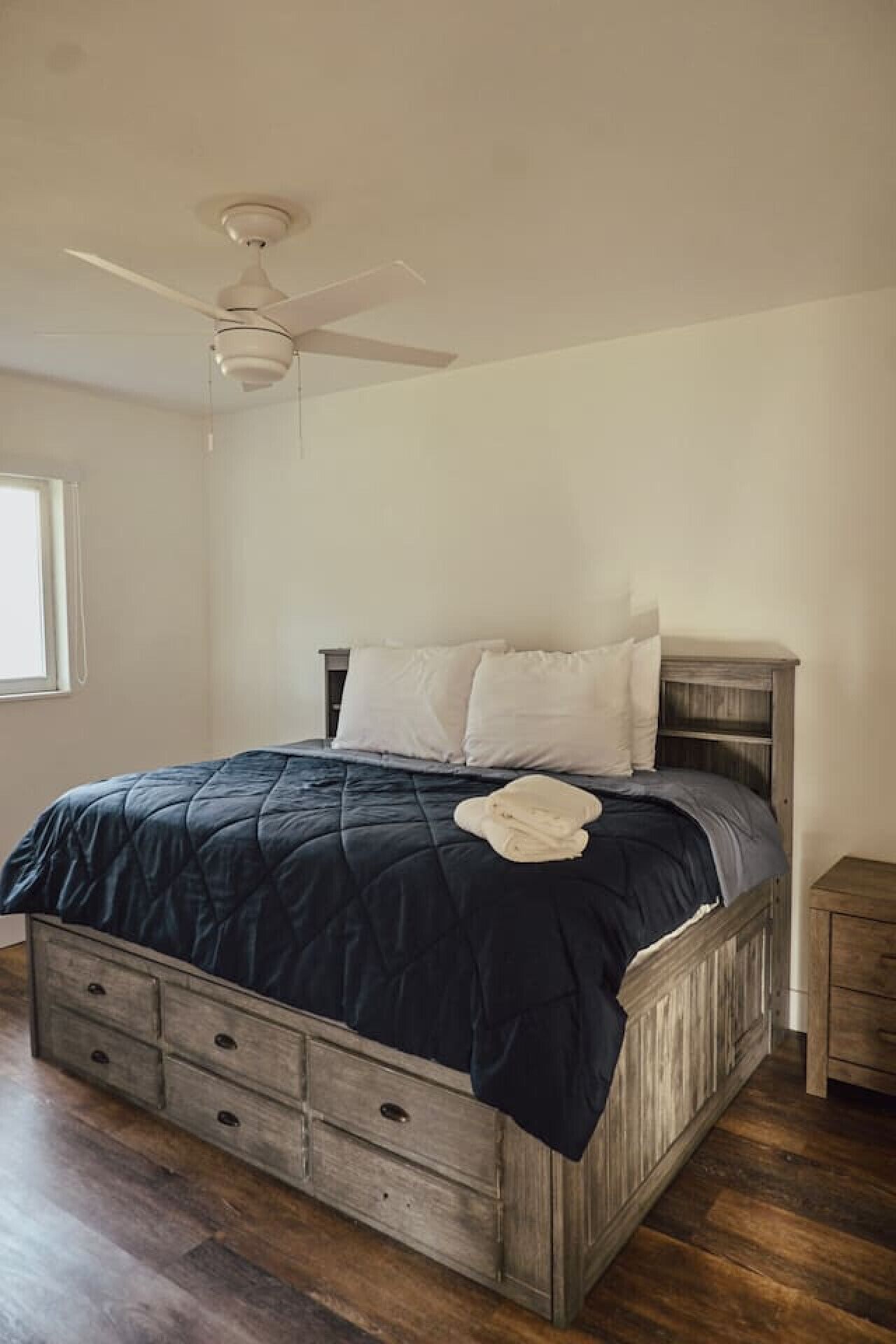 A bedroom with a bed, a ceiling fan, a nightstand, and a window.
