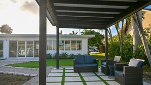 Een overdekt terras met tuinmeubilair, een modern huis op de achtergrond en een verzorgd gazon.