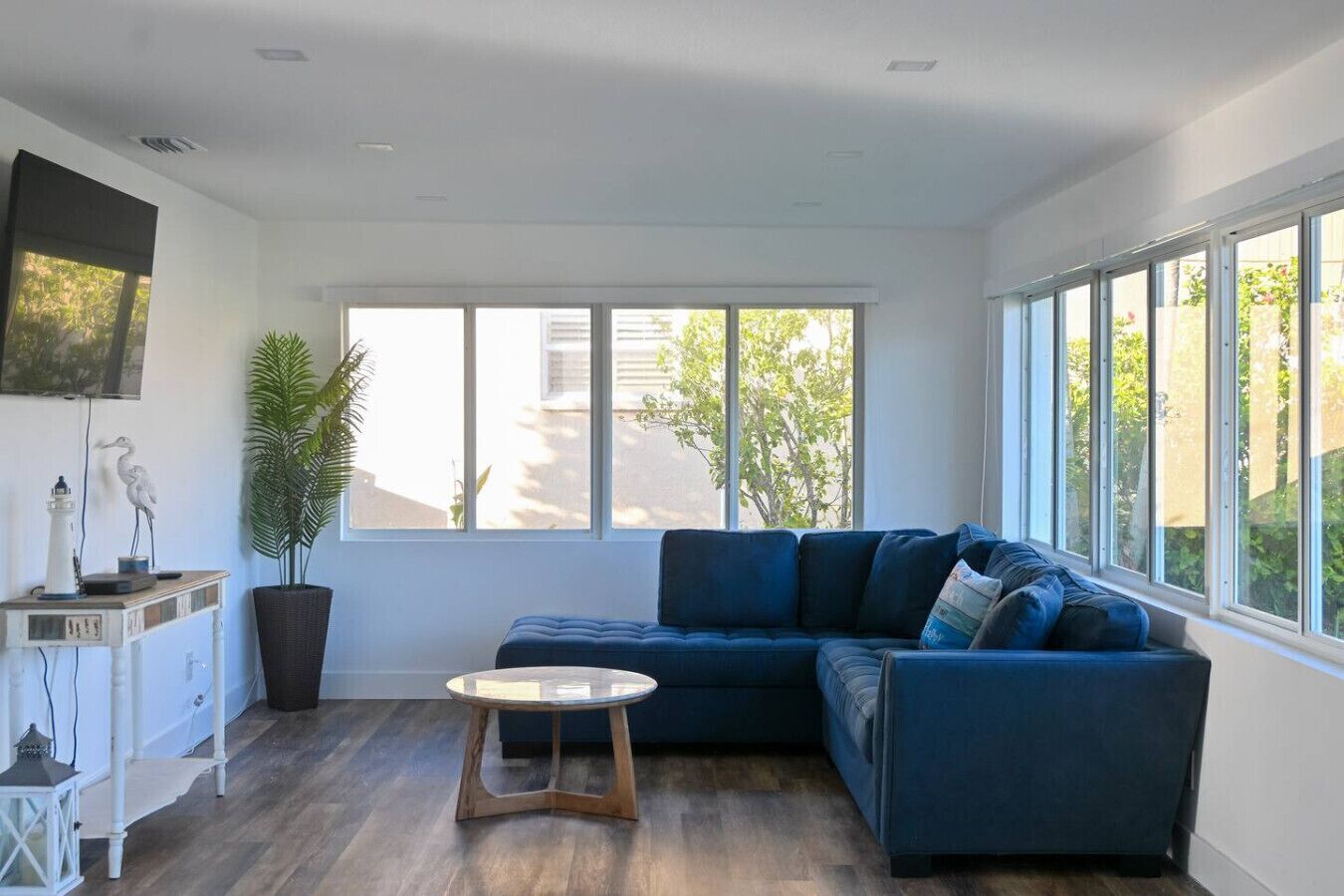 Una sala de estar moderna con un sofá seccional azul oscuro, una mesa de centro redonda de madera y ventanas amplias que dan a vistas de vegetación exterior.