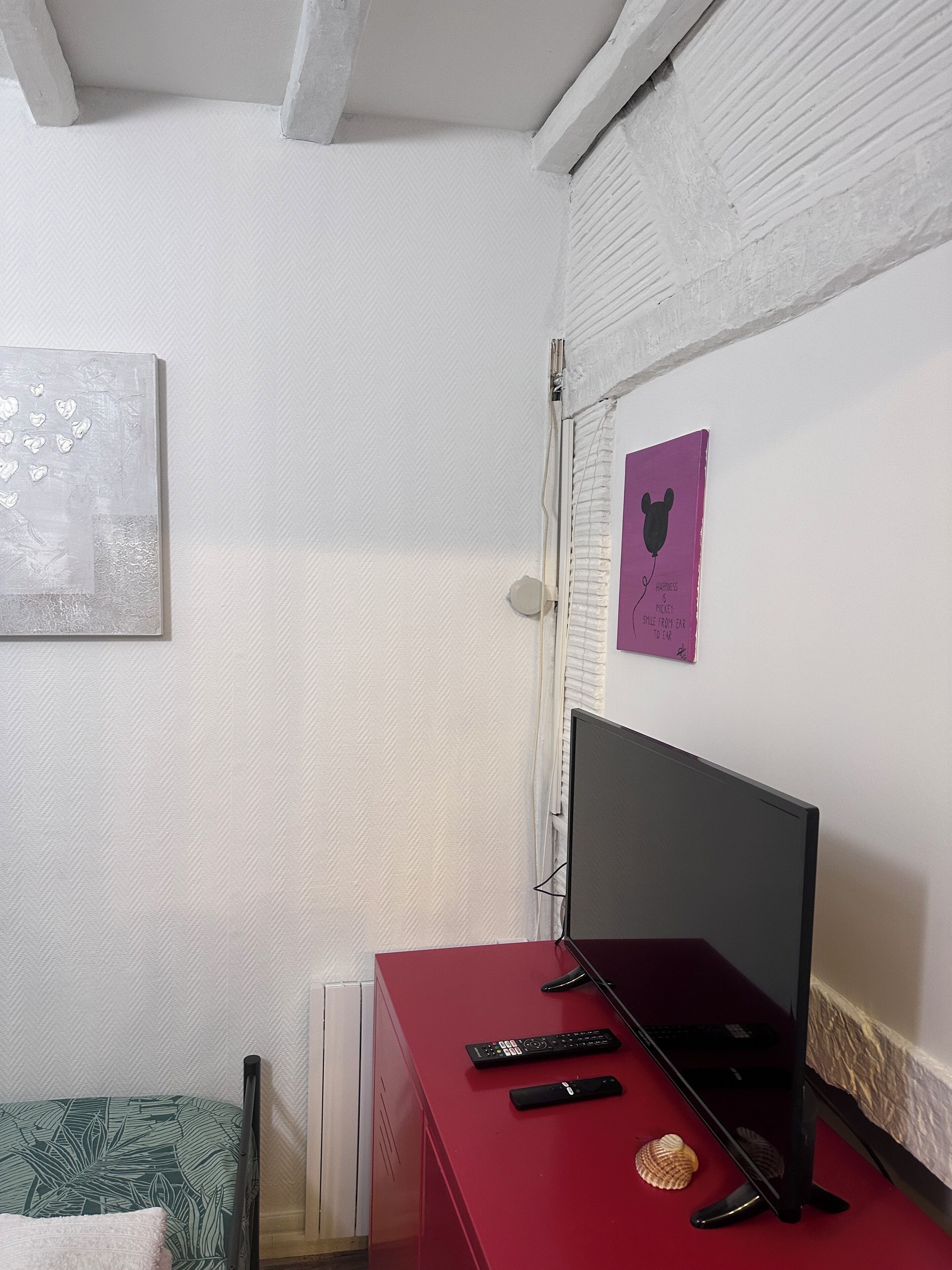 A room with a red desk, a computer monitor, two remote controls, a decorative shell, and a wall with a framed picture.